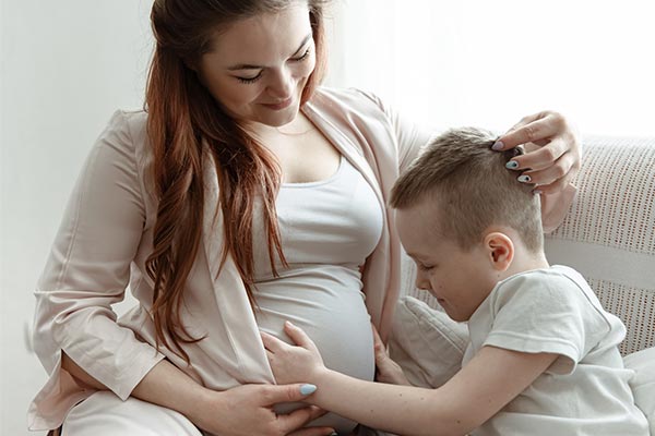 Eine Schwangere sitzt mit einem Kind auf dem Sofa, das Kind umfasst sanft den Babybauch der Frau. Die Frau streicht mit ihrer Hand über den Kopf des Kindes.