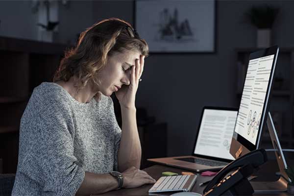 Erschöpft und müde sitzt eine Frau an ihrem dunklen Arbeitsplatz vor dem eingeschalteten Computer.