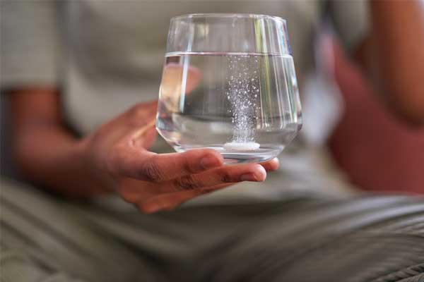 Eine Person hält ein Volles Wasserglas in der Hand, in der sich eine Acetylsalicylsäuretablette löst.