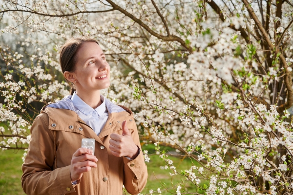 Eine lächelnde Frau steht mit einem Tablettenblister vor einem blühenden Baum.