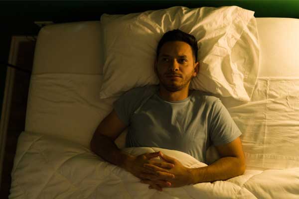 Ein dunkelhaariger Mann, mit verschränkten Händen, liegt im Bett und starrt schlaflos geradeaus. Ein dunkelhaariger Mann, mit verschränkten Händen, liegt im Bett und starrt schlaflos geradeaus.