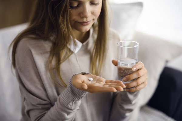 Eine braunhaarige Frau hält in ihrer linken Hand ein Glas mit Wasser und schaut auf eine Naproxentablette in der anderen Hand