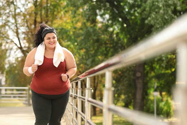 Eine kräftigere Frau in einem dunkelroten Oberteil joggt an einem Geländer vorbei. Eine kräftigere Frau in einem dunkelroten Oberteil joggt an einem Geländer vorbei.