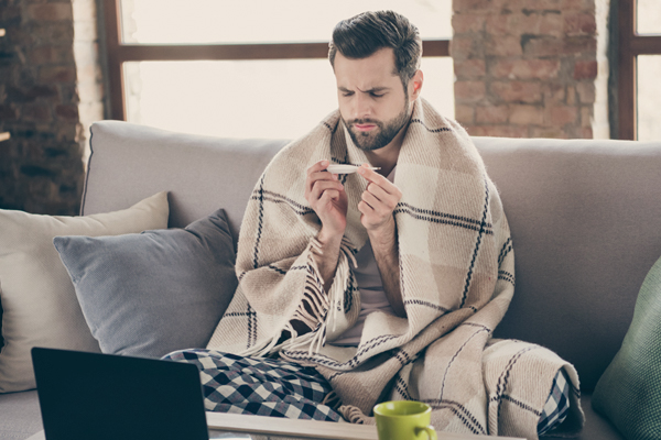 Ein kranker Mann sitzt mit einer Decke um die Schultern und einem Fieberthermometer auf der Couch.