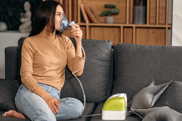 Eine Frau sitzt mit einem Inhaliergerät auf der Couch und inhaliert.