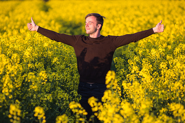 Ein schwarzhaariger Mann mit ausgebreiteten Armen steht in einem Rapsfeld voller Pollen.