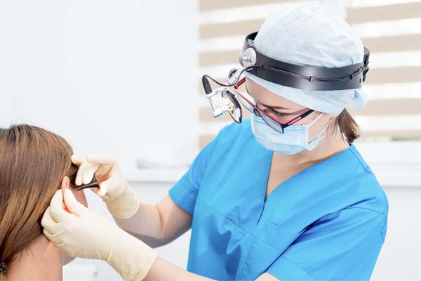 Hals-Nasen-Ohren-Ärztin in einem blauen Ärztekittel untersucht Patient/in am Ohr