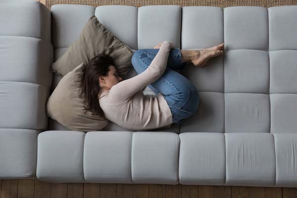 schwarzhaarige Frau liegt in Embryonalstellung auf einem grauen Sofa mit zwei braunen Kissen