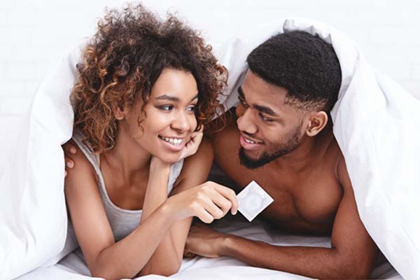 Eine Frau und ein Mann liegen zusammen in einem Bett unter einer Decke und halten ein Kondom in der Hand.