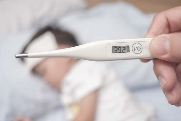 Ein weißes Fieberthermometer, welches 39,2 Grad Celsius anzeigt und im Hintergrund ein dunkelhaariger Junge, der mit einem weißen Tuch auf der Stirn im Bett liegt.