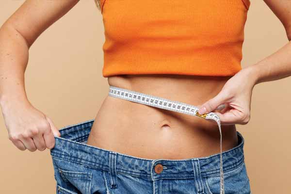 Eine Frau misst ihren flachen Bauch mit einem Maßband in einer zu weiten Jeans als Symbol für erfolgreiches Abnehmen und das Ankurbeln der Fettverbrennung. Eine Frau misst ihren flachen Bauch mit einem Maßband in einer zu weiten Jeans als Symbol für erfolgreiches Abnehmen und das Ankurbeln der Fettverbrennung.
