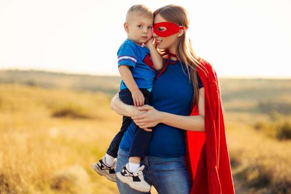 Eine junge Mutter im Superhelden-Kostüm hält ihr Kleinkind auf dem Arm.