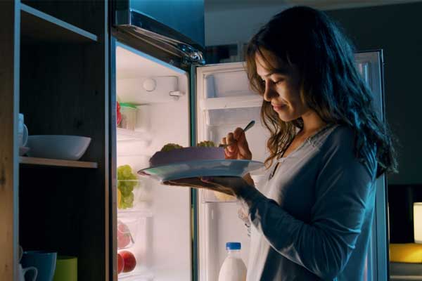 Eine Frau isst wegen einer Heißhungerattacke einen Kuchen vor dem Kühlschrank.