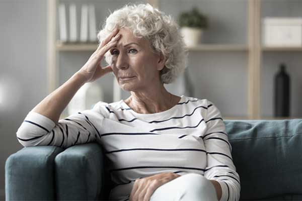 Eine ältere Frau sitzt auf einem Sofa und hält sich aufgrund einer Migräneattacke die Stirn.