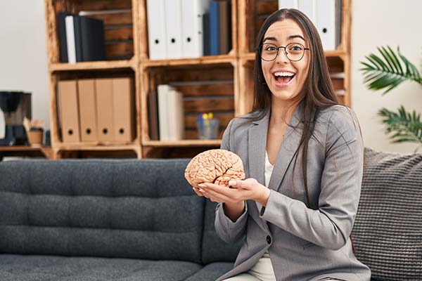 Eine junge Frau mit einem Blazer und Brille sitzt lächelnd auf einen grauen Sofa und hält ein Gehirn als Modell in der Hand. Eine junge Frau mit einem Blazer und Brille sitzt lächelnd auf einen grauen Sofa und hält ein Gehirn als Modell in der Hand.