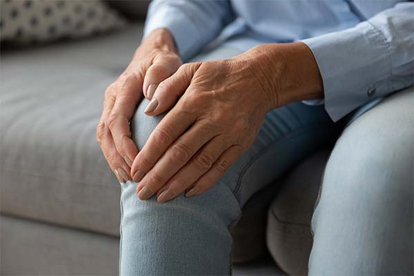 Hände einer älteren Person umfassen ein schmerzendes Knie beim Sitzen auf dem Sofa.