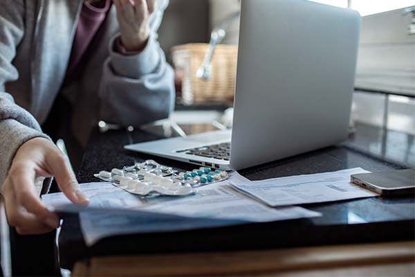 Eine Person sichtet am Laptop Dokumente, die neben mehreren Blisterpackungen mit Medikamenten und einem Smartphone auf einem Tisch liegen. Eine Person sichtet am Laptop Dokumente, die neben mehreren Blisterpackungen mit Medikamenten und einem Smartphone auf einem Tisch liegen.