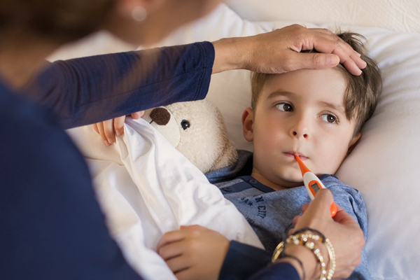 Eine Mama misst ihrem Jungen mit einem Fieberthermometer Fieber im Mund. Dabei legt sie ihm eine Hand auf die Stirn.
